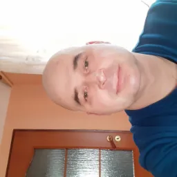 Uśmiechnięty mężczyzna w niebieskiej bluzie na tle pomarańczowej ściany i drewnianych drzwi z szybą. Widoczny fragment białej zasłony.