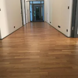 Długi korytarz z nowo ułożonym, jasnym parkietem drewnianym w jodełkę, widoczne drzwi i białe ściany z czarnymi akcentami.