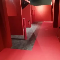 Czerwone, minimalistyczne wnętrze z labiryntem korytarzy, wykończone matową farbą, z widocznymi elementami instalacji elektrycznej i dokumentami na postumencie.