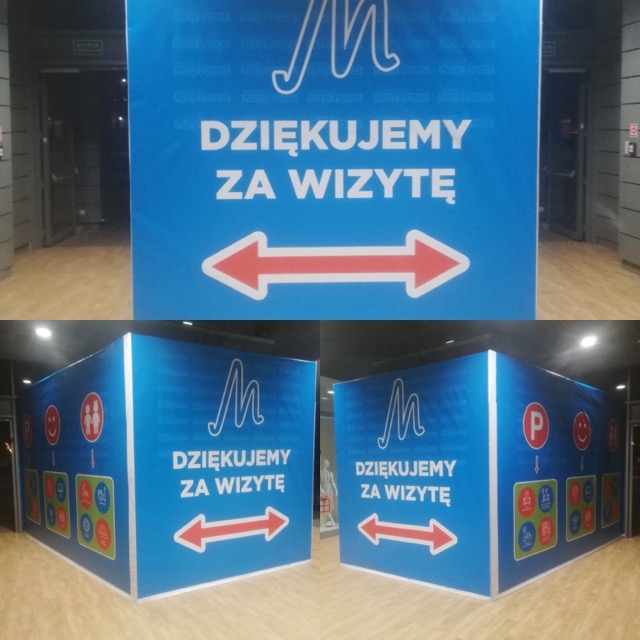 Duży, niebieski, przestrzenny napis informacyjny z logo i strzałką wskazującą kierunek, z tekstem 'Dziękujemy za wizytę' umieszczony wewnątrz budynku.