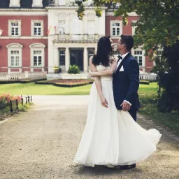 Para narzeczeńska w objęciach przed malowniczym budynkiem w Czarnogoździcach; panna młoda w zwiewnej sukni, pan młody w granatowym smokingu, widoczny park i starodawna architektura w tle.