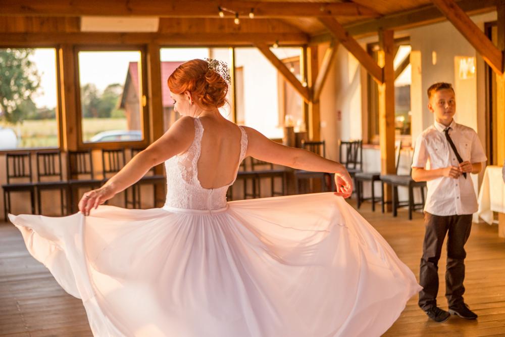 Kobieta w białej sukni z odkrytymi plecami, rozkładająca spódnicę, w tle chłopiec w koszuli i krawacie oraz okna z widokiem na zewnątrz.