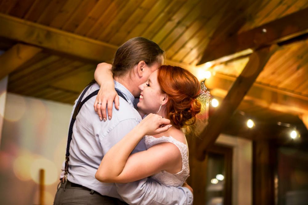 Para młoda w objęciach podczas pierwszego tańca, ona w białej sukni z koronkową górą i upiętymi rudymi włosami z ozdobą, on w jasnej koszuli z szelkami, drewniany sufit z żarówkami w tle.