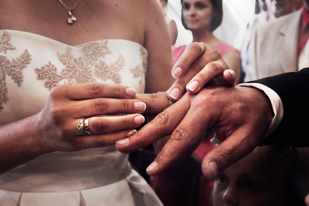 Panna młoda wkłada obrączkę na palec pana młodego, widoczne dłonie, fragment sukni ślubnej z koronkowymi aplikacjami, w tle goście weselni.