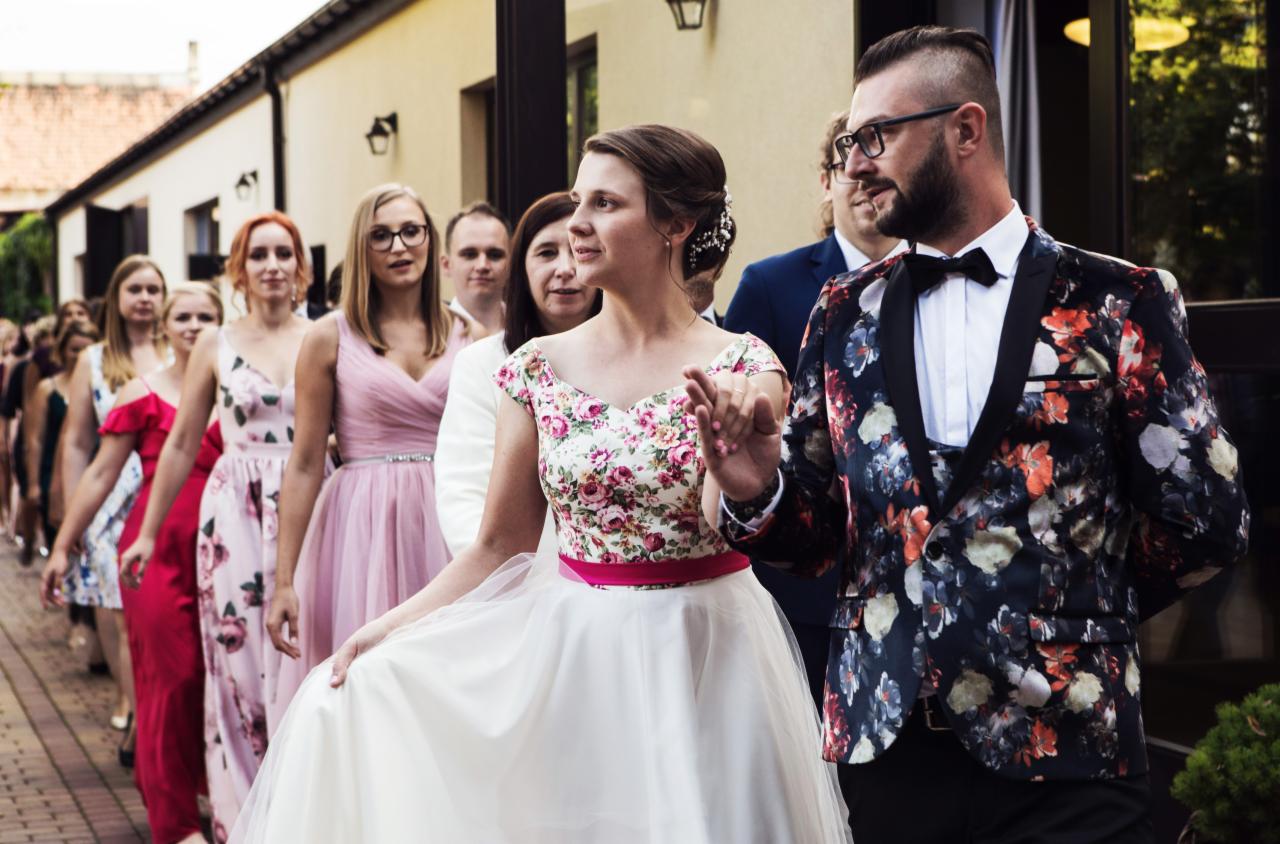 Para młoda i druhny w sukniach balowych, idący w rzędzie przed budynkiem, mężczyzna w kwiecistym smokingu.