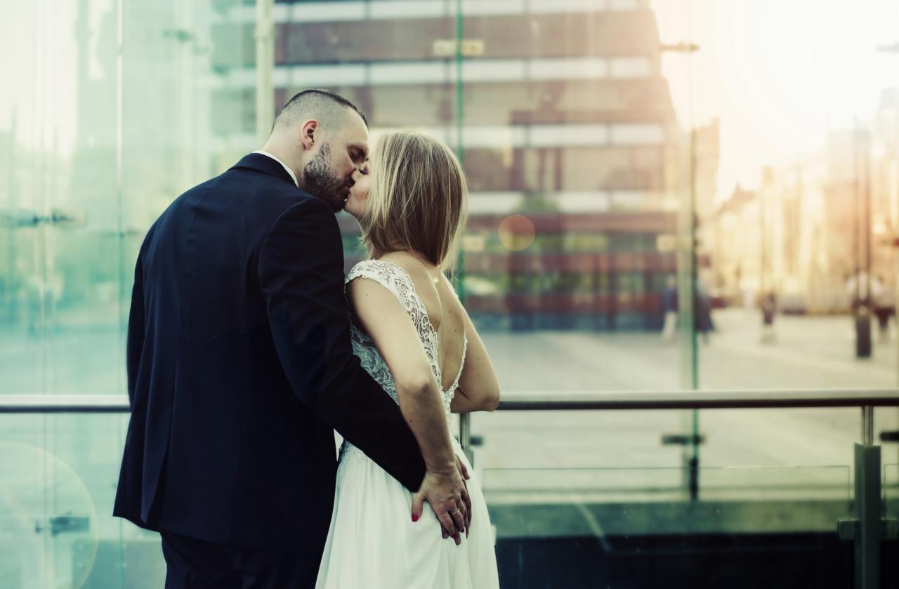 Para całująca się na tle szklanej fasady budynku w miejskim krajobrazie, kobieta w białej sukni z koronkową górą, mężczyzna w czarnym garniturze, delikatne promienie słońca w tle.