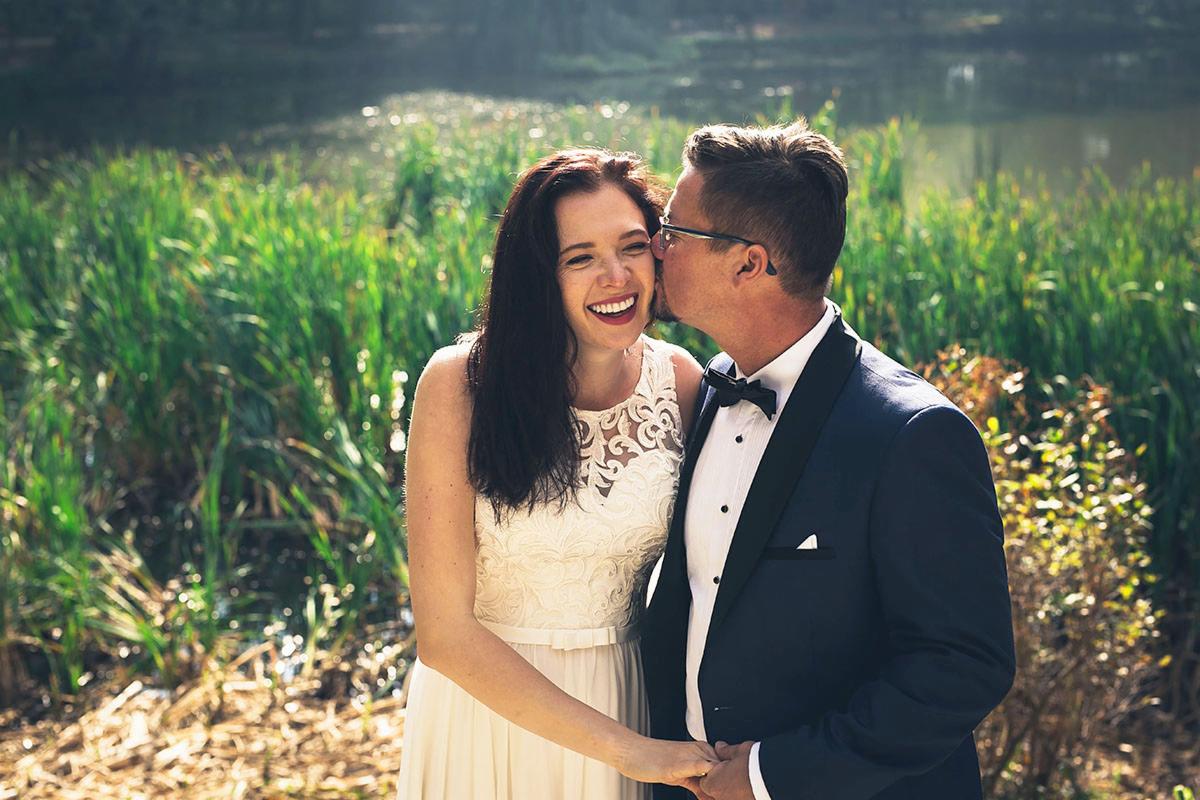 Uśmiechnięta panna młoda w koronkowej sukni, całowana w policzek przez pana młodego w granatowym smokingu z czarną muchą, na tle jeziora i zieleni.