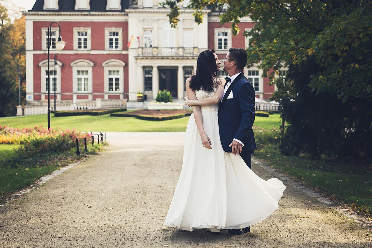 Para narzeczeńska w objęciach przed malowniczym budynkiem w Czarnogoździcach; panna młoda w zwiewnej sukni, pan młody w granatowym smokingu, widoczny park i starodawna architektura w tle.