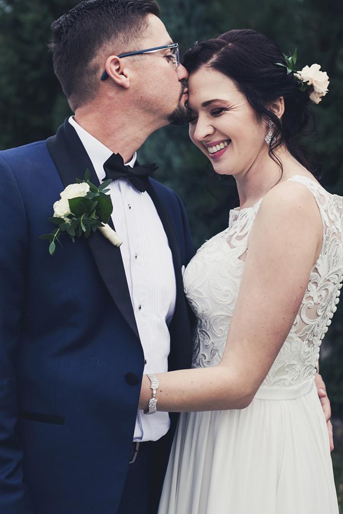 Mężczyzna w granatowym smokingu z butonierką całuje kobietę w koronkowej sukni ślubnej z kwiatami we włosach.