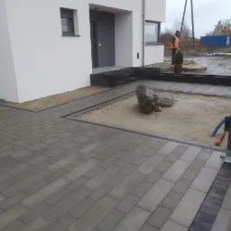 Ułożona szara kostka brukowa przed nowoczesnym domem z białymi ścianami, widoczny fragment ogrodzenia, piaskowe podłoże z dekoracyjnymi kamieniami i małym drzewkiem, pracownik w odblaskowej...