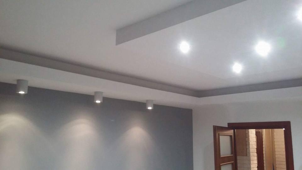 Nowoczesne wnętrze: biały sufit z podwieszanymi elementami i punktowym oświetleniem, szara ściana z trzema lampami, otwarte drewniane drzwi w tle.