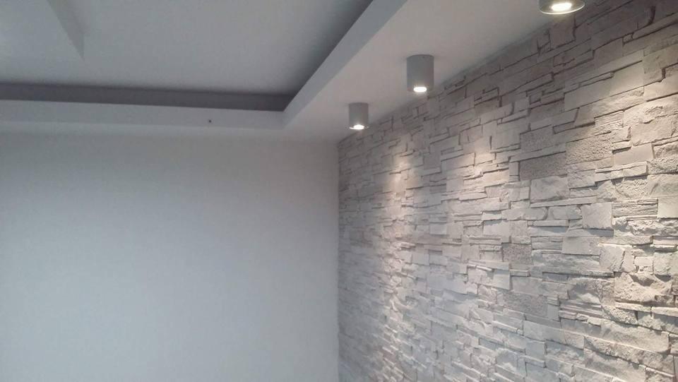 Ściana wykończona dekoracyjną okładziną z imitacji kamienia, oświetlona trzema punktowymi lampami sufitowymi, sąsiaduje z gładką, pomalowaną ścianą i podwieszanym sufitem.