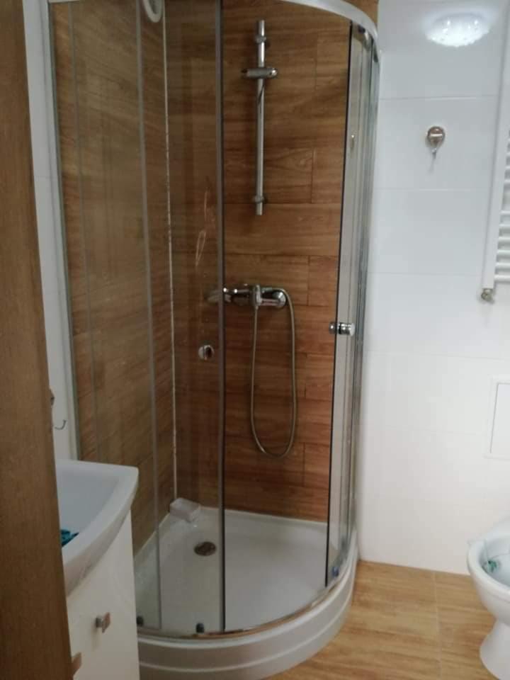 Kabina prysznicowa z drewnopodobnymi płytkami ściennymi, okrągła kabina z chromowanymi elementami, widoczna umywalka i toaleta w tle, jasne oświetlenie.