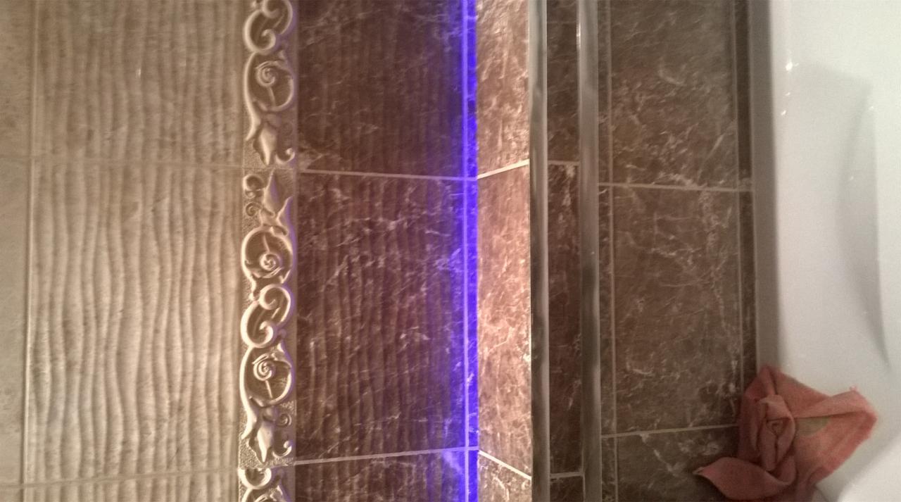 Fragment łazienki z dekoracyjnymi, beżowymi i brązowymi płytkami, podświetlonym narożnikiem prysznica i ręcznikiem. Płytki w dwóch wzorach: jeden z ornamentami kwiatowymi, drugi z fakturą...