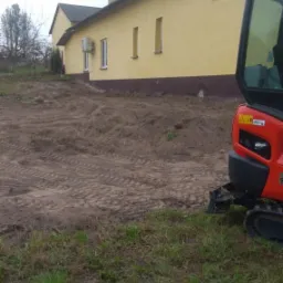 Częściowo zniwelowany teren przed żółtym budynkiem, widoczny fragment mini koparki Kubota z prawej strony.