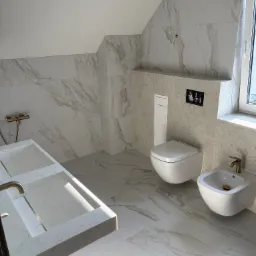 Jasna łazienka na poddaszu z marmurowymi płytkami, wiszącą toaletą i bidetem. Umywalka z dwoma komorami i złotą armaturą. Widoczne okno i skos dachu.