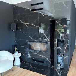Grestom - Elegancki salon z kominkiem obłożonym czarnym marmurem z złotymi żyłkami, drewniana podłoga, biały fotel, czarne ściany, nowoczesny design.