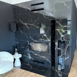 Elegancki salon z kominkiem obłożonym czarnym marmurem z złotymi żyłkami, drewniana podłoga, biały fotel, czarne ściany, nowoczesny design.