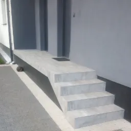 Nowoczesne, szare schody wejściowe wyłożone płytkami ceramicznymi, z piłką do koszykówki leżącą pod spodem, na tle szarej elewacji budynku.