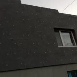 Ściana budynku pokryta ciemnoszarym styropianem, przymocowanym kołkami. Widoczne okno z białymi ramami oraz fragment dolnej części elewacji pokrytej szarym tynkiem.