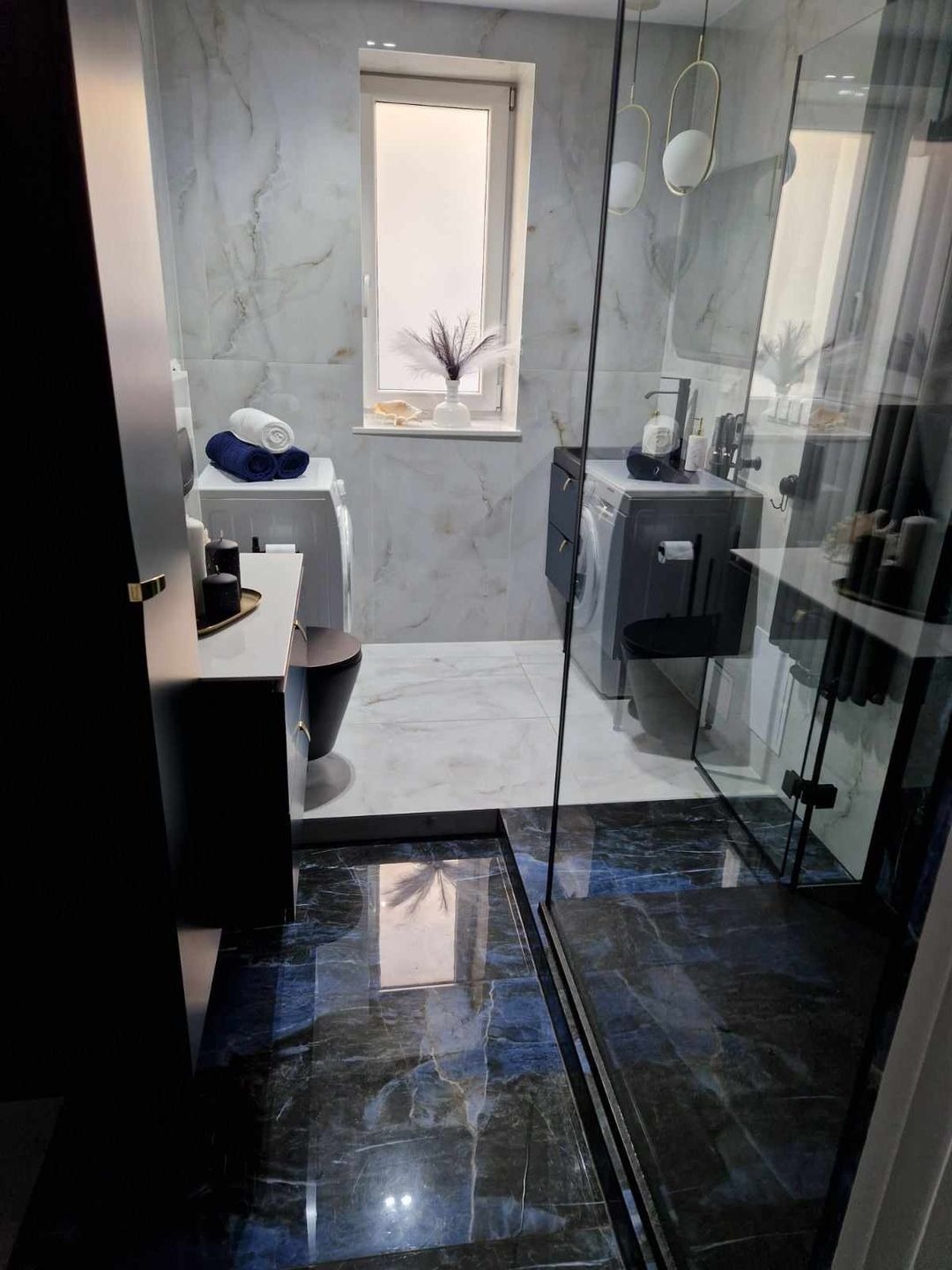 Nowoczesna łazienka z marmurowymi płytkami w odcieniach bieli i szarości, z akcentami czerni w armaturze i meblach, oraz niebieską mozaiką na podłodze w strefie prysznica, oddzieloną szklaną kabiną.