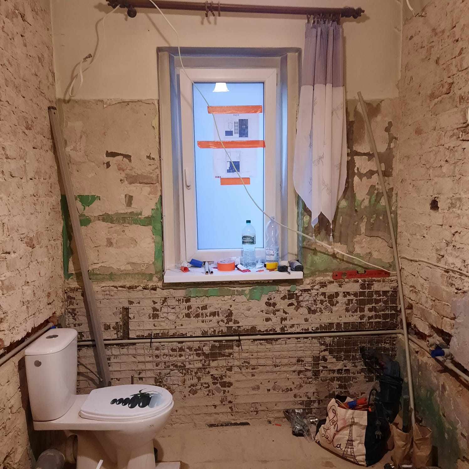 Łazienka w trakcie prac przygotowawczych: odsłonięta cegła, widoczne instalacje, okno z planami, narzędzia i materiały budowlane, toaleta z naklejką na desce.
