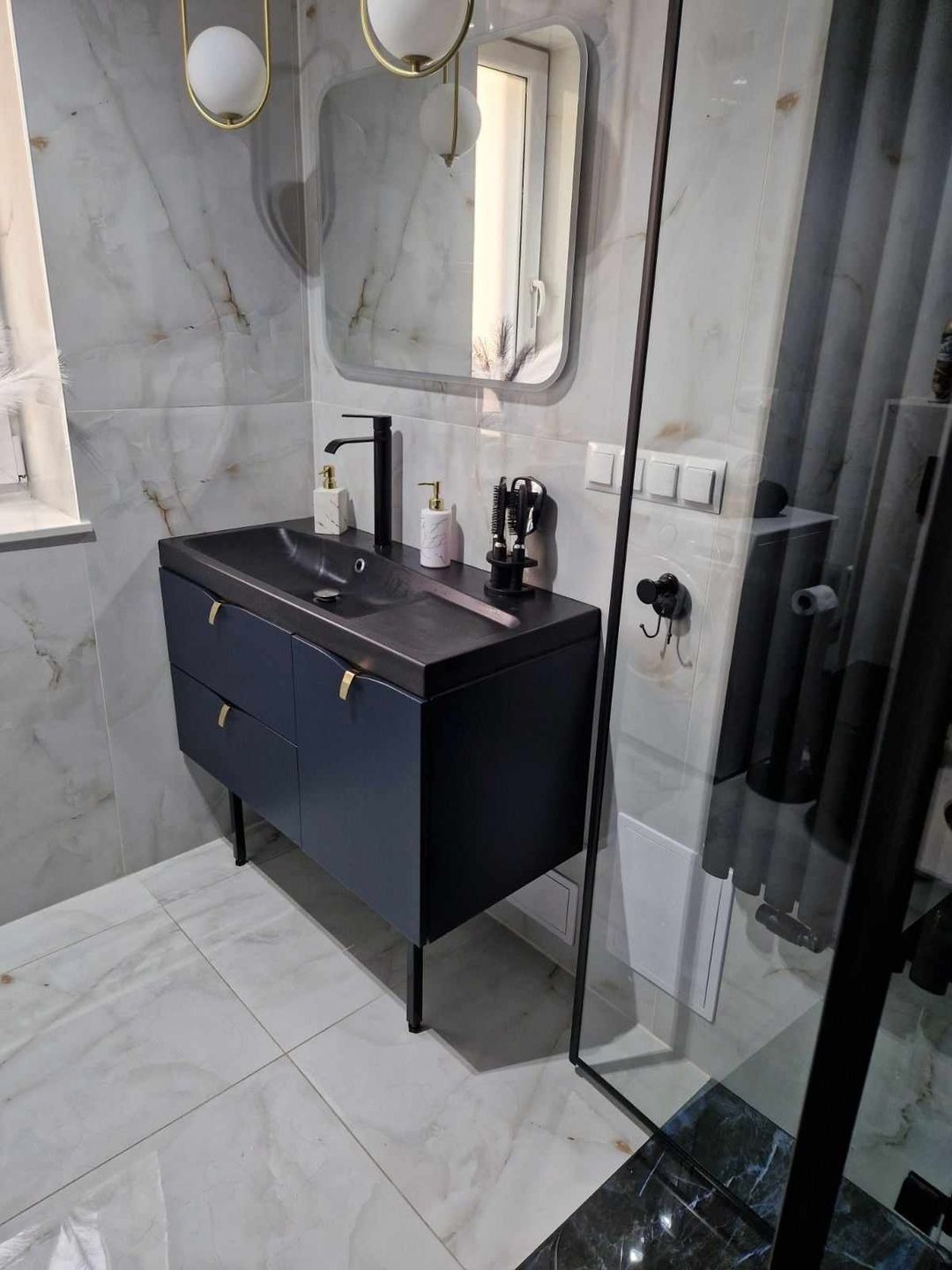 Nowoczesna łazienka z umywalką nablatową, czarną armaturą i szafką w kolorze granatowym, marmurowe płytki na ścianach i podłodze, oświetlenie z kulistymi lampami.