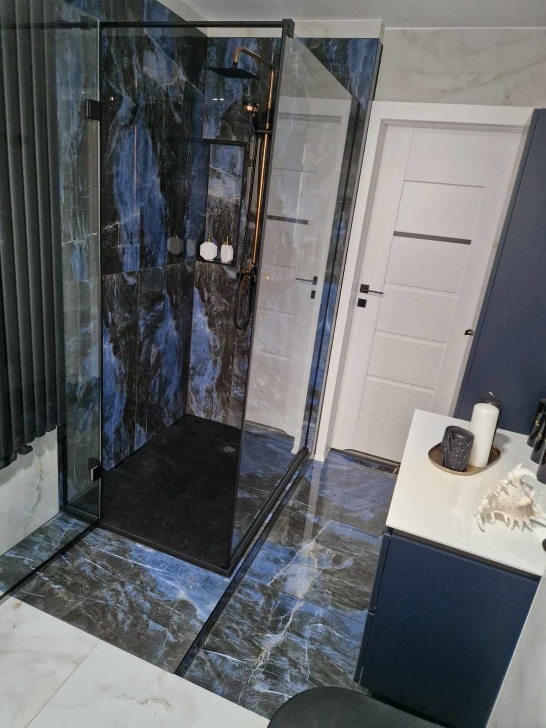 Nowoczesna łazienka z kabiną prysznicową typu walk-in, wykończona płytkami imitującymi marmur w odcieniach granatu i błękitu, z czarną armaturą i minimalistyczną umywalką z dekoracjami.