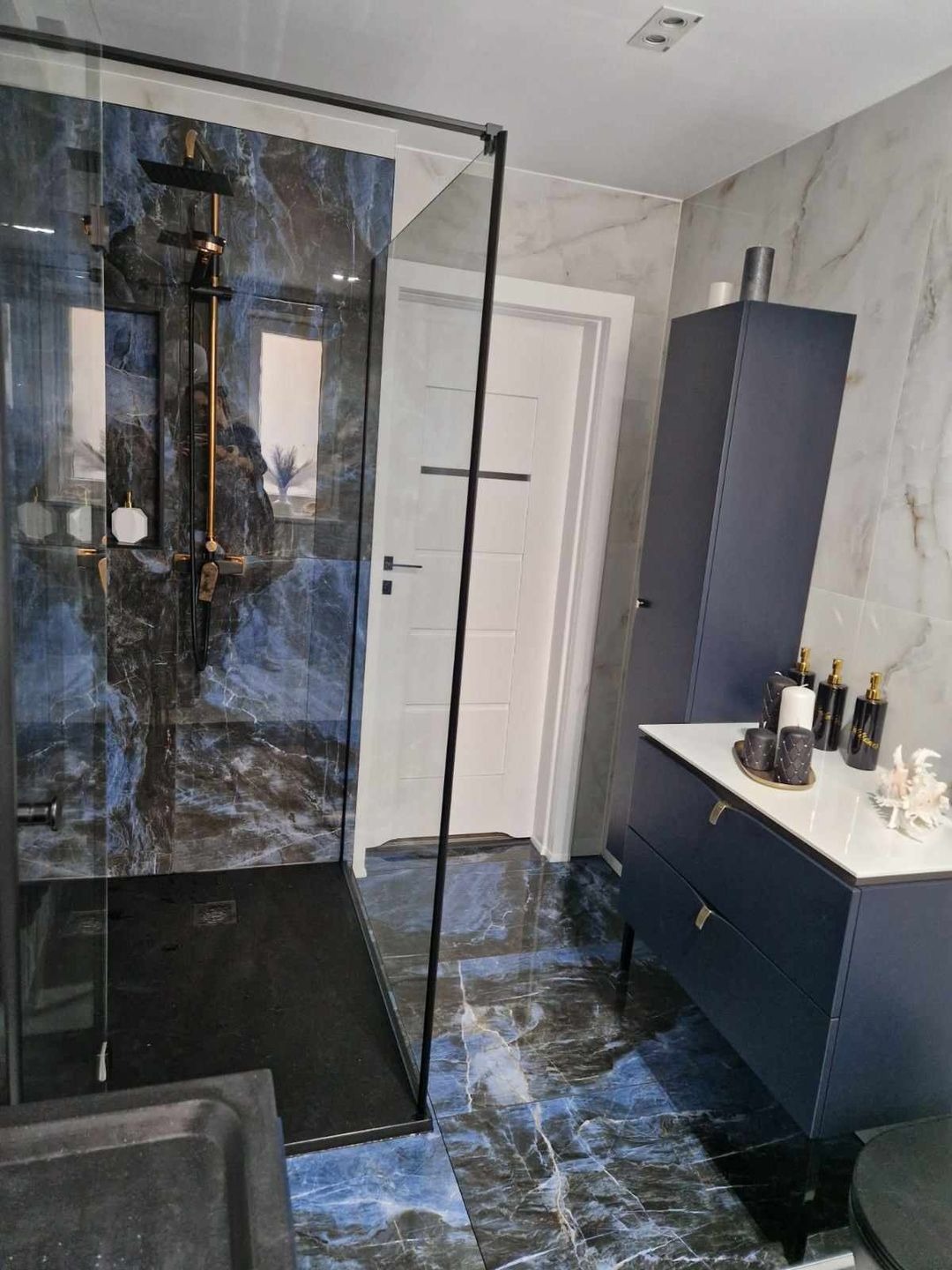Nowoczesna łazienka z kabiną prysznicową typu walk-in, wykończona płytkami imitującymi marmur w odcieniach granatu i szarości, z czarną armaturą i szafką z umywalką w kolorze granatowym.