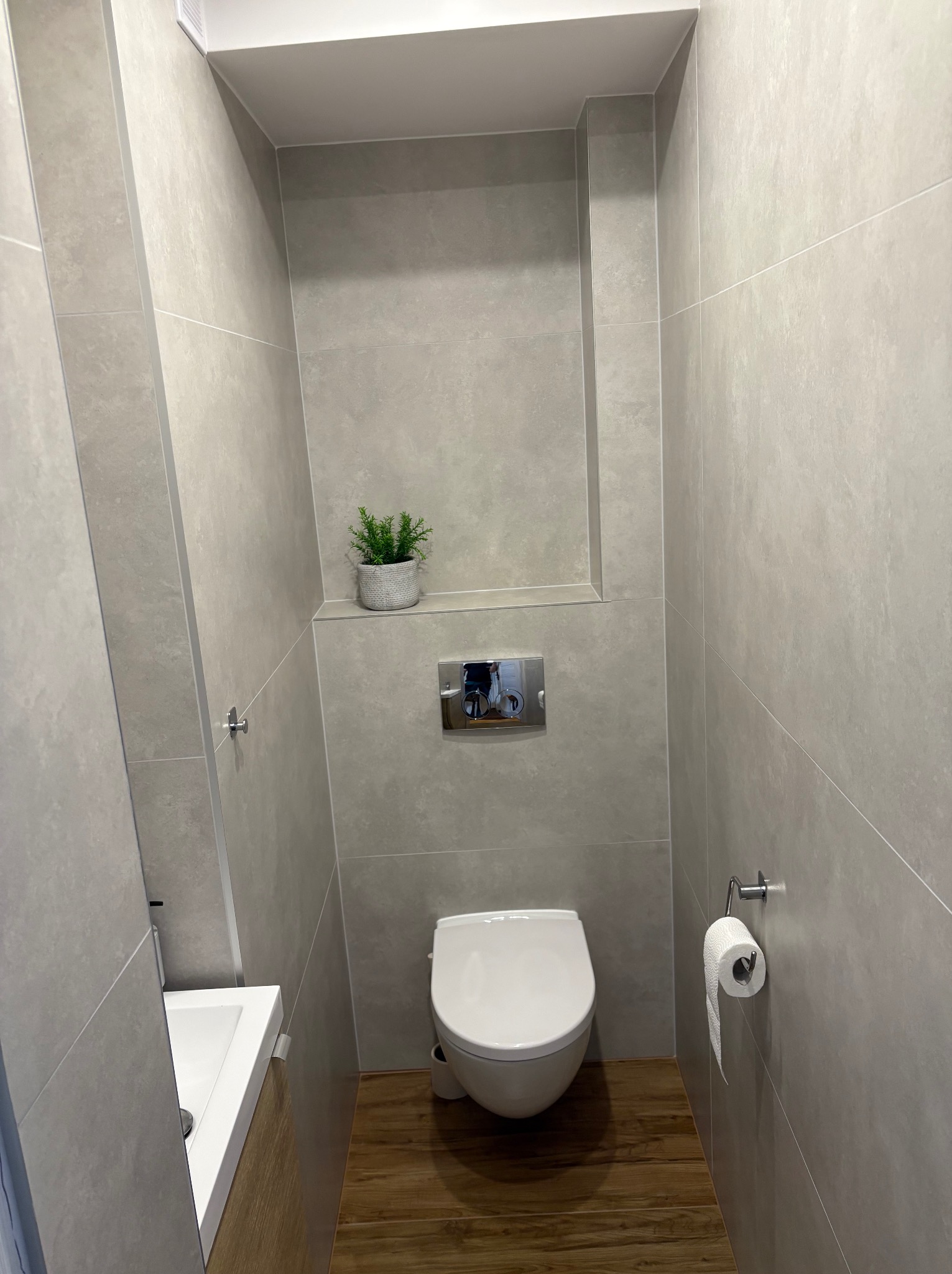 Nowoczesna, minimalistyczna toaleta wykończona szarymi płytkami, z podwieszaną miską WC, drewnopodobną podłogą, małą umywalką z szafką, rośliną w doniczce we wnęce i chromowanym uchwytem na papier...