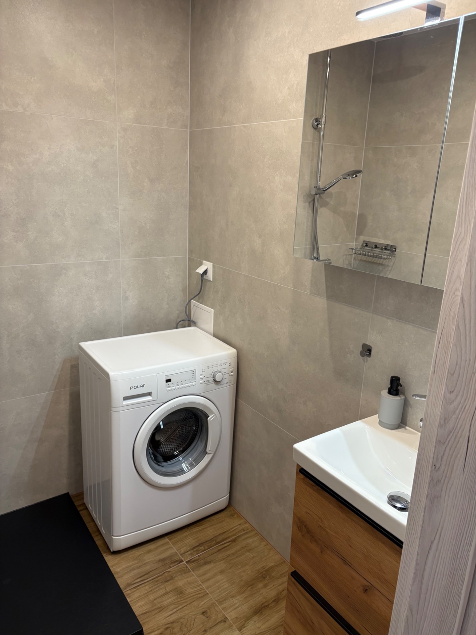 Nowoczesna łazienka z beżowymi płytkami, białą pralką Polar, umywalką z szafką z drewnopodobnym frontem i dużym lustrem z oświetleniem nad nim.
