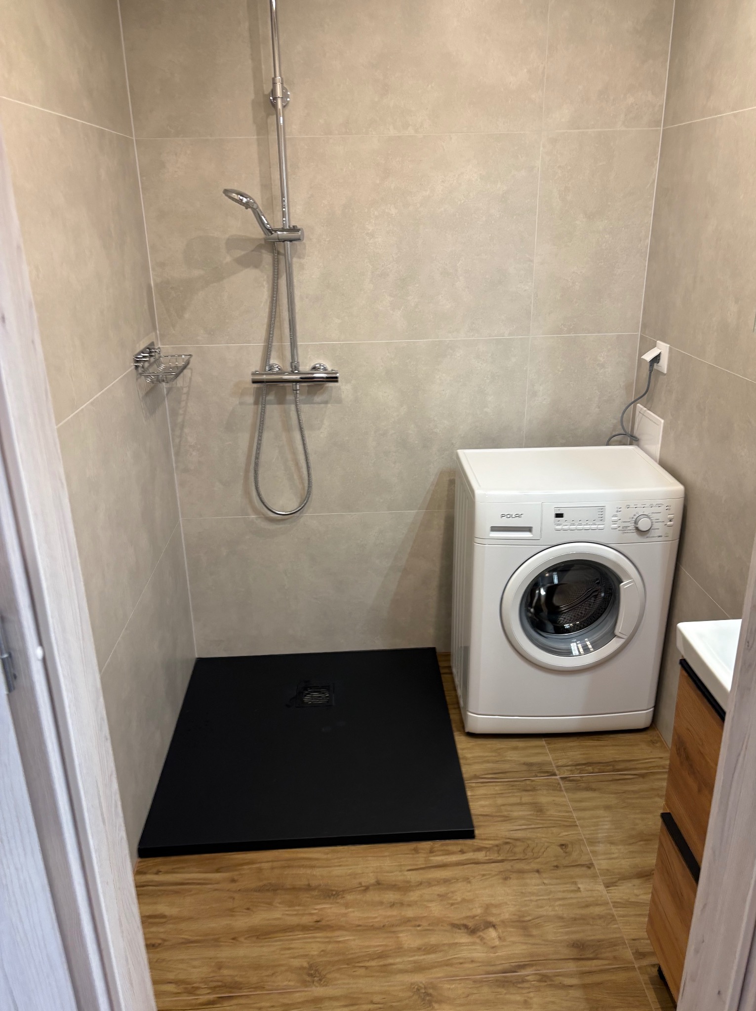 Wykończona łazienka w Lędzinach z kabiną prysznicową z czarnym brodzikiem, pralką i drewnopodobnymi płytkami na podłodze.
