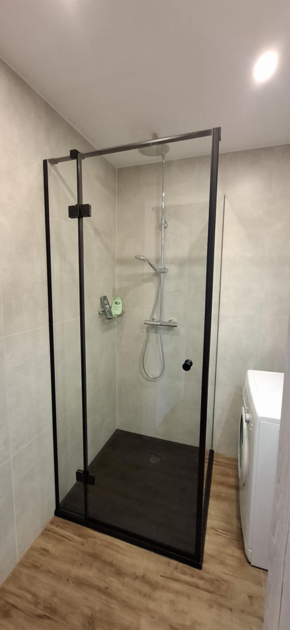 Kabina prysznicowa z czarną ramą na tle beżowych płytek, z widoczną pralką z boku i drewnopodobną podłogą.