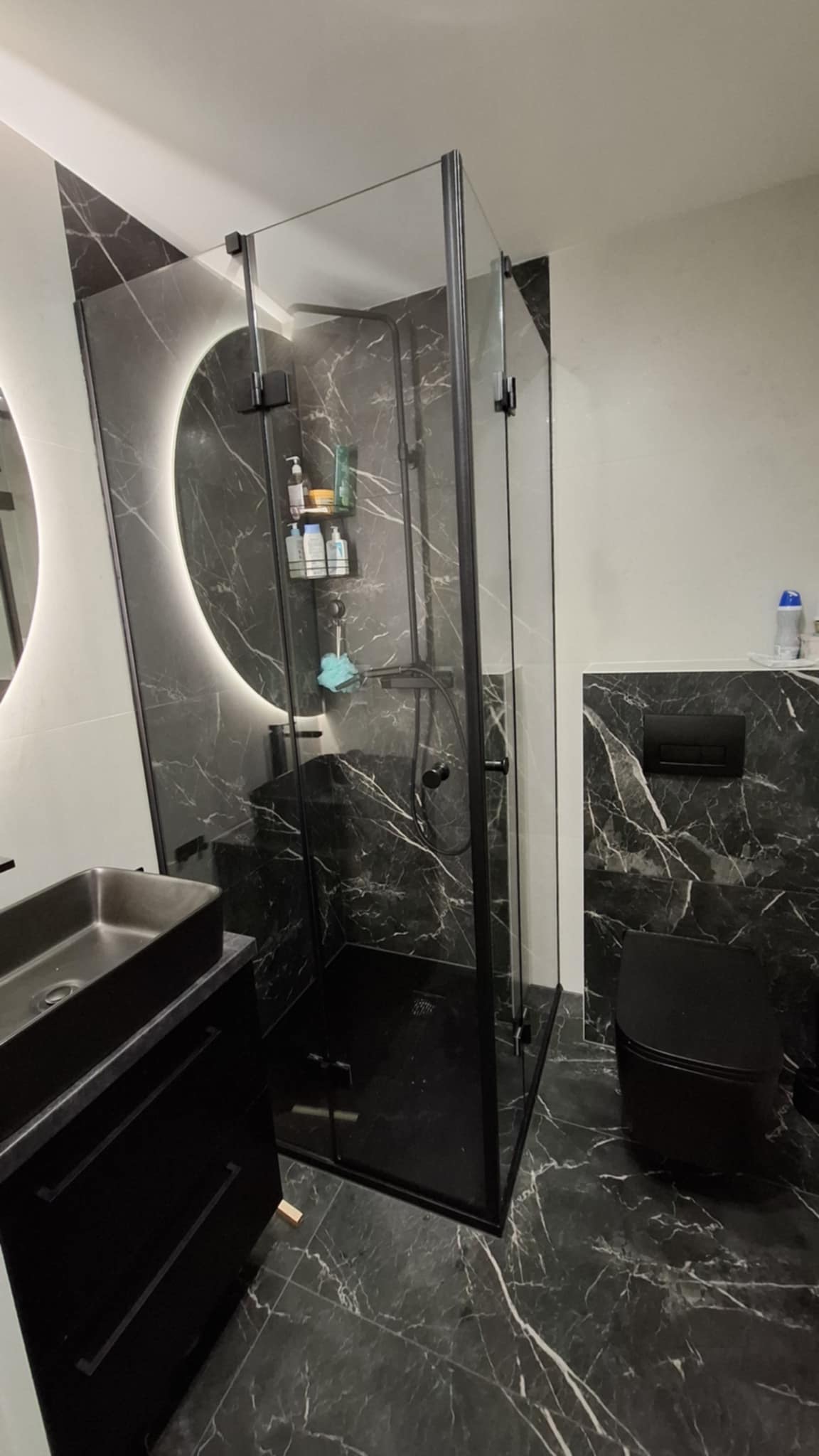 Nowoczesna łazienka z czarnymi marmurowymi płytkami, kabiną prysznicową typu walk-in z czarnymi ramami, czarną umywalką nablatową i podświetlanym lustrem LED