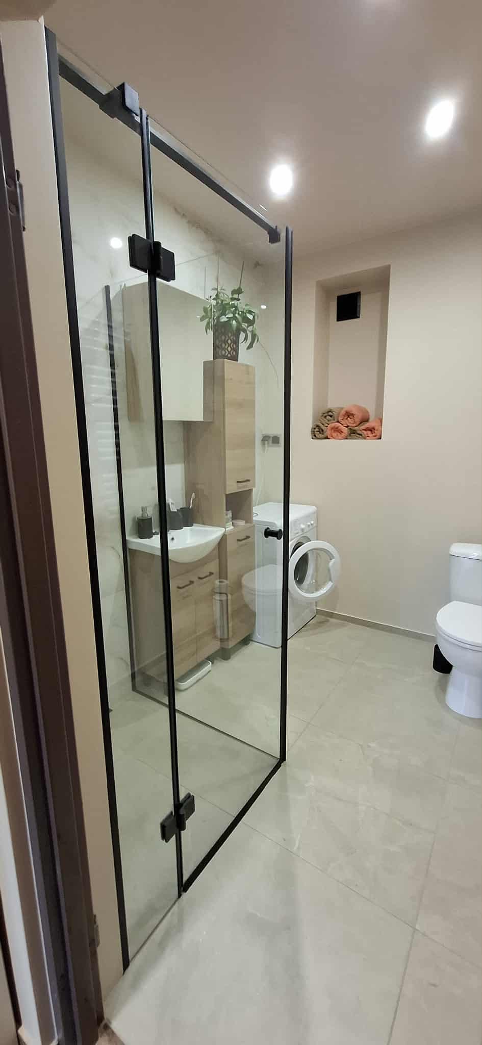 Nowoczesna łazienka z kabiną prysznicową typu walk-in z czarnymi profilami, umywalką z szafką z jasnego drewna i pralką. Wnęka z ręcznikami dodaje uroku.