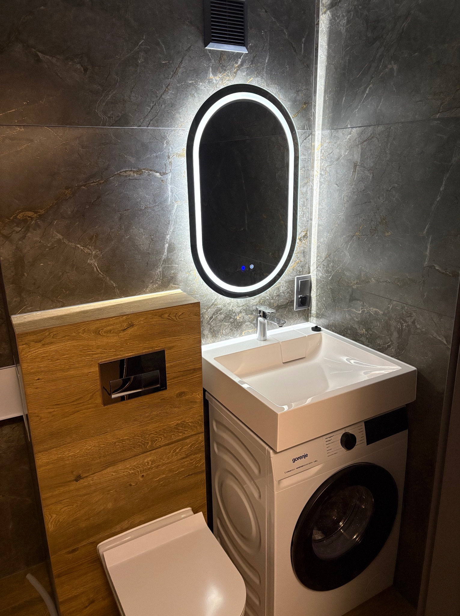 Nowoczesna łazienka z umywalką nad pralką, podświetlanym lustrem i marmurowymi płytkami. Drewniana obudowa WC z chromowanym przyciskiem. Minimalistyczny design.