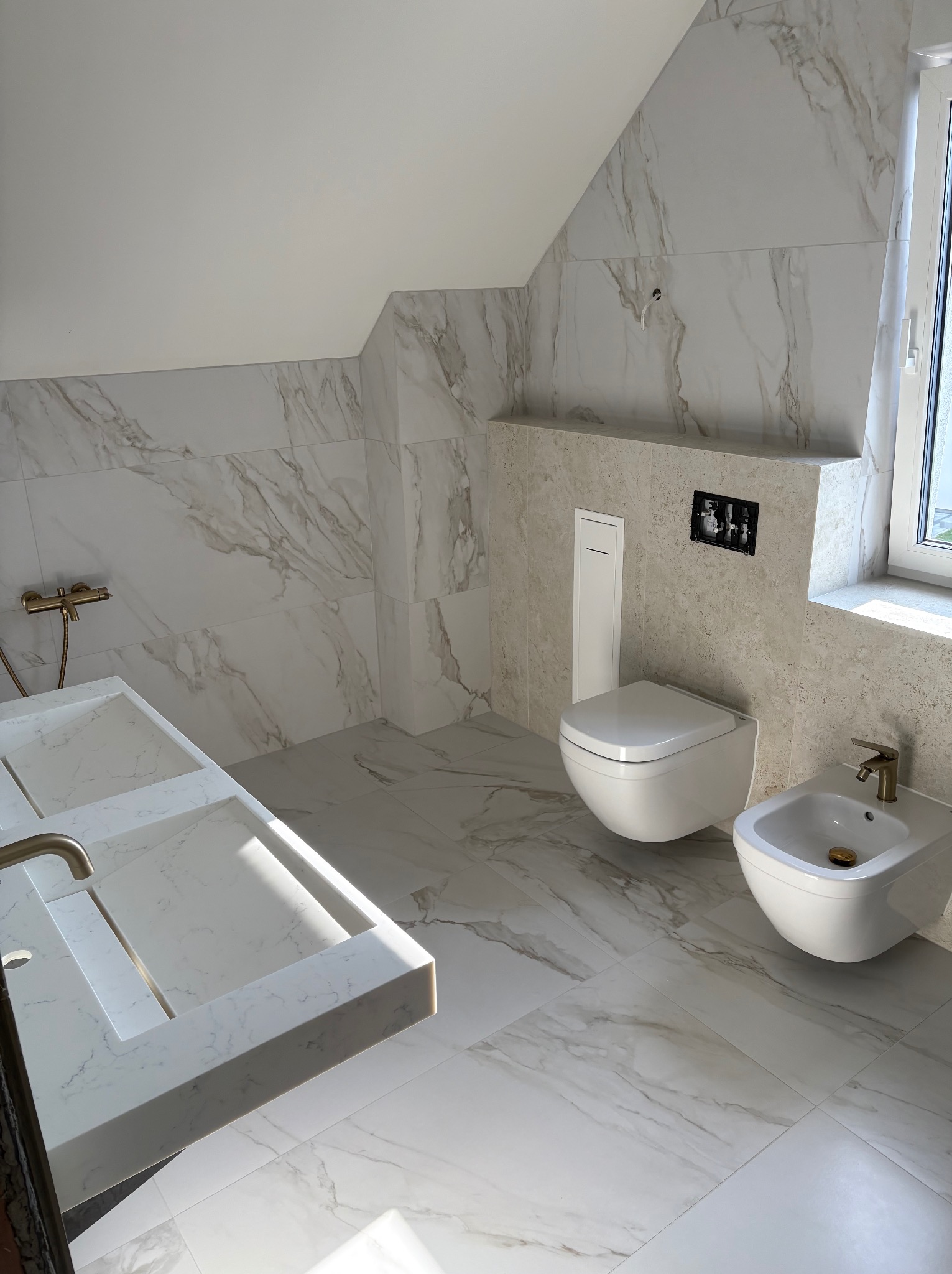 Jasna łazienka na poddaszu z marmurowymi płytkami, wiszącą toaletą i bidetem. Umywalka z dwoma komorami i złotą armaturą. Widoczne okno i skos dachu.