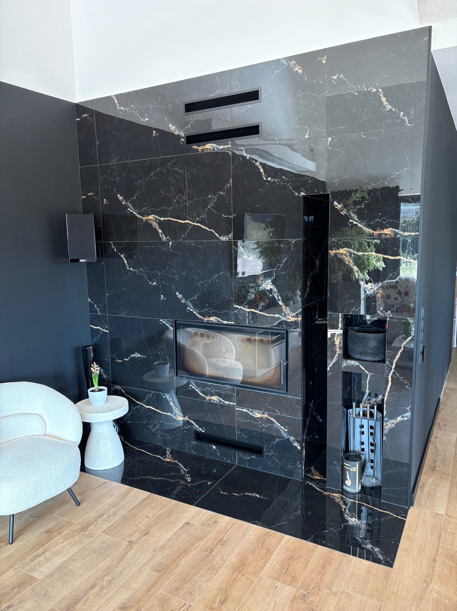 Elegancki salon z kominkiem obłożonym czarnym marmurem z złotymi żyłkami, drewniana podłoga, biały fotel, czarne ściany, nowoczesny design.