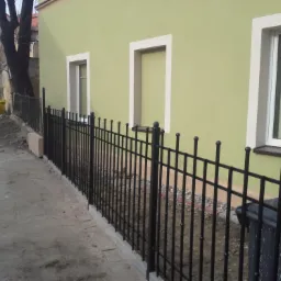 Nowo postawione, czarne, metalowe ogrodzenie posesji z kulkami na szczytach, na tle świeżo pomalowanej na jasnozielono ściany budynku z białymi obramowaniami okien.