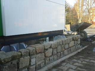 Niska, kamienna ścianka oporowa z nieregularnych bloków skalnych, zabezpieczająca biały pawilon, z widocznym fragmentem chodnika i taczką w tle.