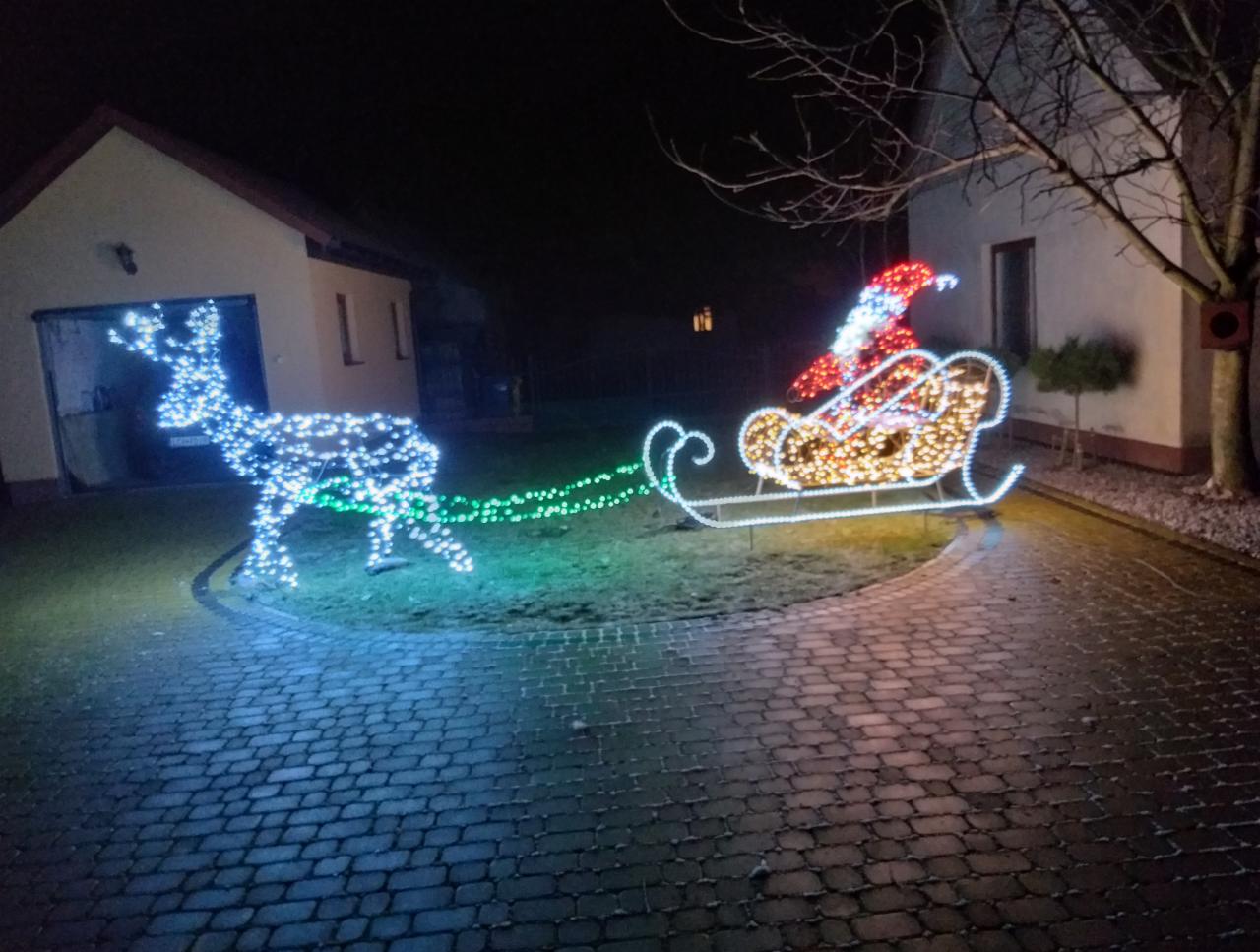 Świąteczna dekoracja ogrodowa: podświetlany renifer ciągnący sanie z Mikołajem, wykonane z białych i kolorowych lampek LED na tle domu i kostki brukowej.