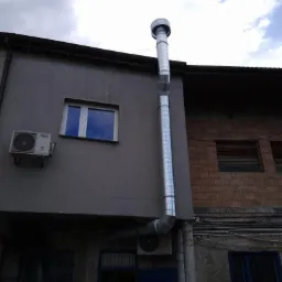 Zewnętrzny, metalowy przewód wentylacyjny zamontowany na ścianie budynku z widocznym oknem, klimatyzatorem i drabiną opartą o wejście.