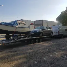 Ł&oacute;dź pod plandeką i samoch&oacute;d osobowy przewożone na lawecie ciągniętej przez busa na tle budynk&oacute;w i drzew.