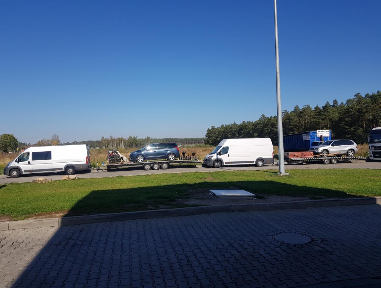 Dwa białe busy i dwie lawety z samochodami osobowymi, w tle las i błękitne niebo, widok w słoneczny dzień.