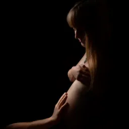 Ciemna, klimatyczna fotografia ciążowa z profilu, ukazująca kobietę z długimi blond włosami i delikatnym makijażem, osłaniającą piersi rękami, z dłonią partnera na brzuchu, na czarnym tle.