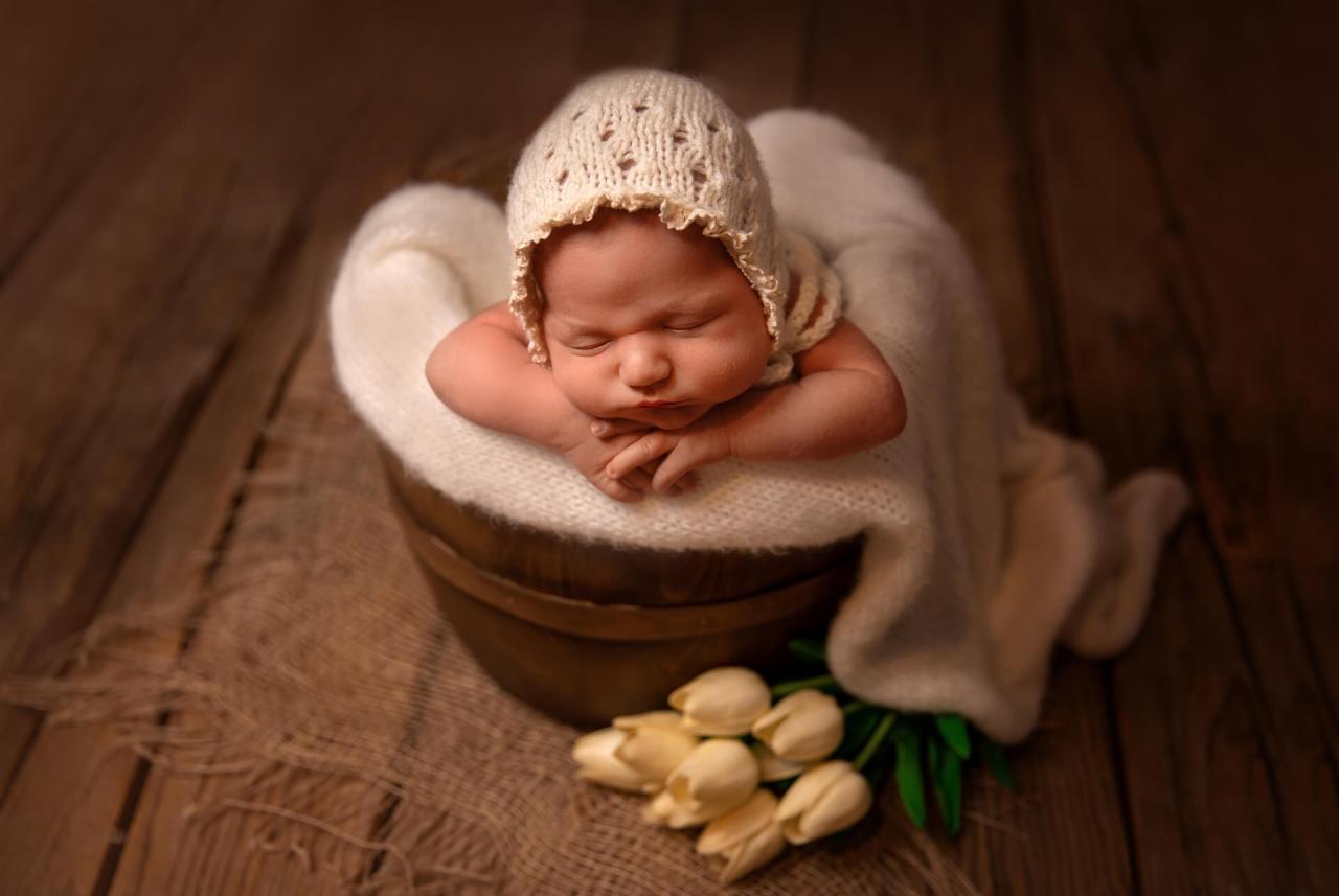 Śpiący noworodek w ażurowym czepku, ułożony w drewnianym wiaderku wyłożonym białym kocykiem, z bukietem kremowych tulipanów u podstawy.
