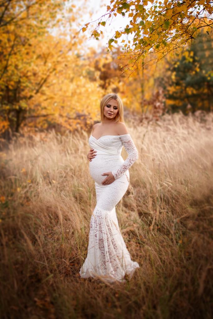 Kobieta w ciąży w białej, koronkowej sukni pozuje na łące z wysoką trawą w jesiennej scenerii.