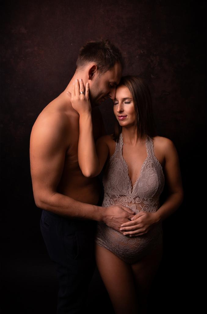 Czułe ujęcie pary: mężczyzna obejmuje kobietę w koronkowej bieliźnie, dłonie na jej brzuchu, sesja ciążowa w studio na ciemnym tle.