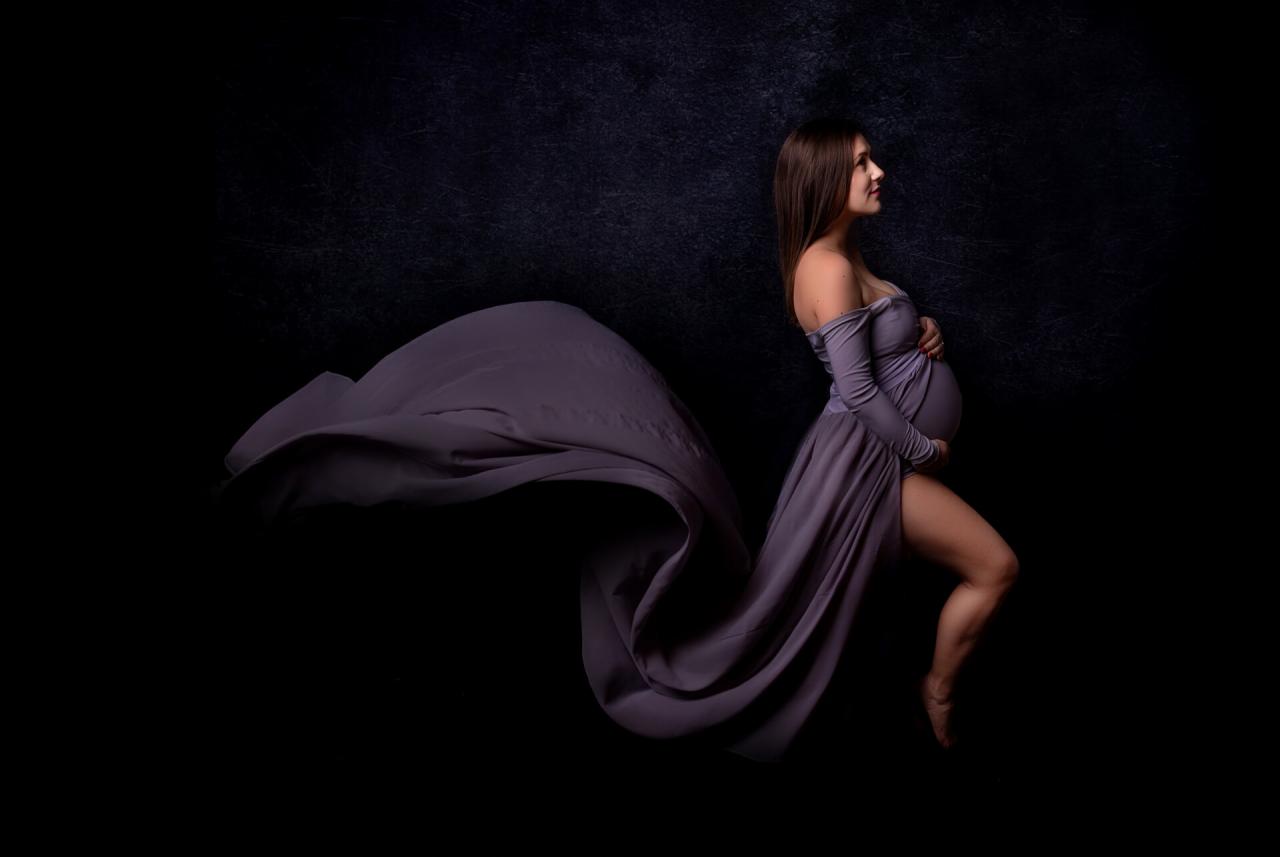 Kobieta w ciąży pozuje w studio na ciemnym tle, ubrana w powłóczystą, liliową suknię z rozcięciem, materiał sukni tworzy efektowny tren.