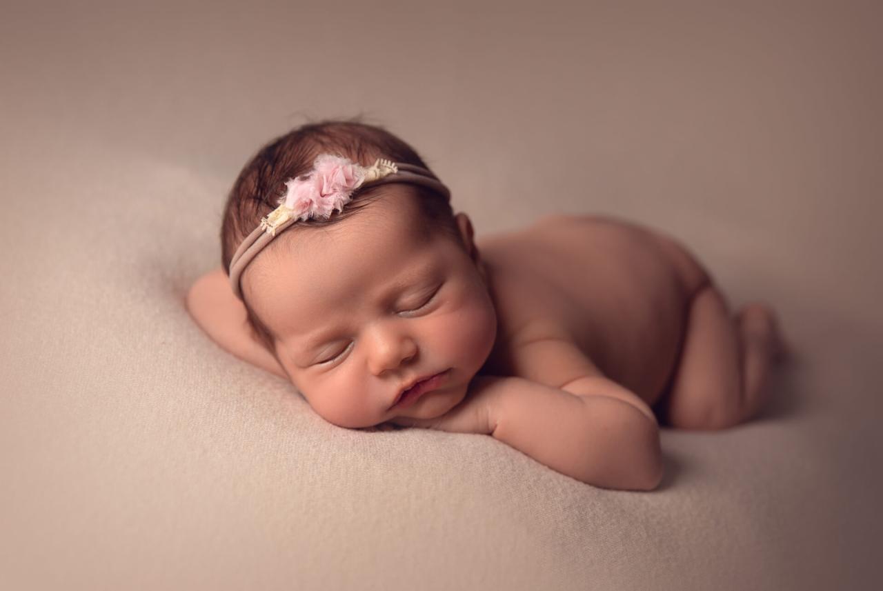 Śpiąca noworodka z różową opaską na głowie, leżąca na beżowym tle, delikatne oświetlenie.